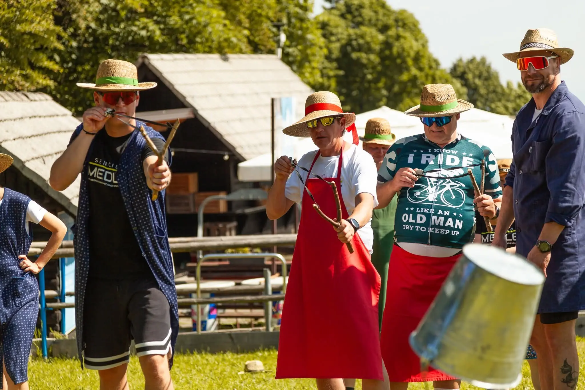 Skupina ljudi na prostem tekmuje s fračami, vsi nosijo klobuke in sončna očala.