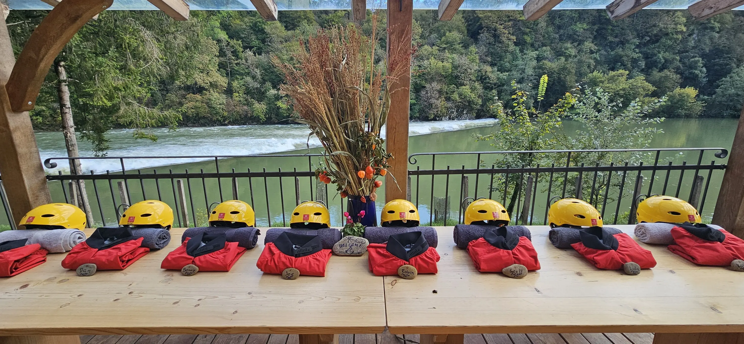 Miza z rumenimi čeladami, brisačami in rdečimi oblekami ob reki v naravnem okolju.