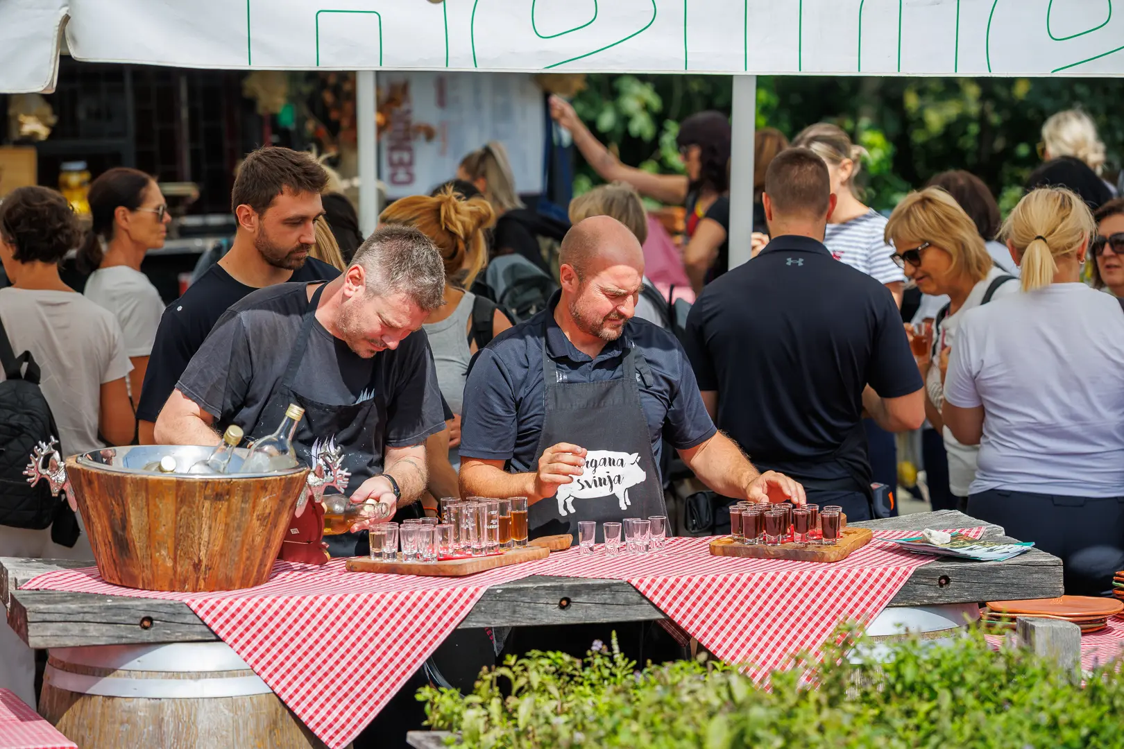 Moška pripravljata pijače na stojnici z rdečimi karirastimi prti v množici ljudi na festivalu.