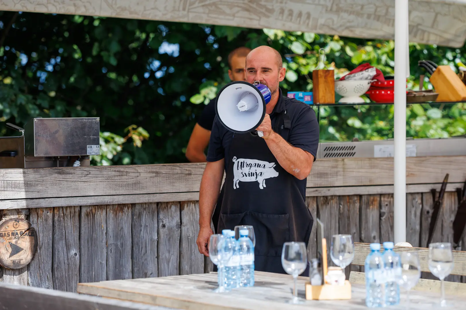 Moški z megafonom oblečen v predpasnik, stoji za leseno mizo z vodo in kozarci