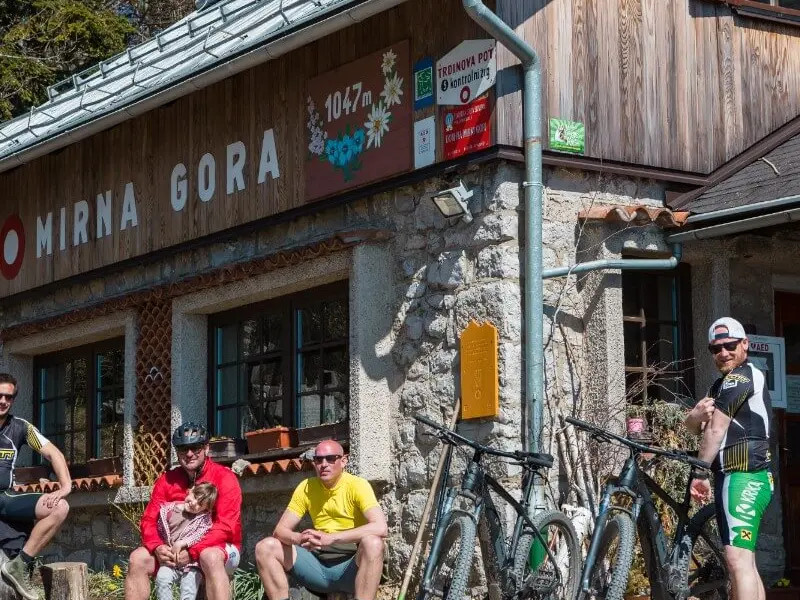 Skupina kolesarjev pred gorskim zavetiščem Mirna gora na sončen dan.